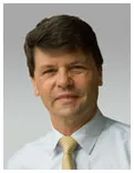 Brian Pittwood, smiling, in a pale blue shirt and gold tie, against a grey backdrop