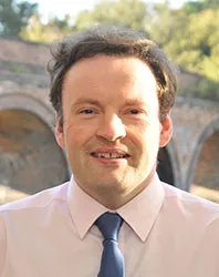 Mark Robinson, smiling, in a white shirt and navy tie, brick arches behind