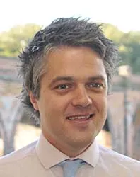 Phil Polden, smiling, in a pale pink shirt and blue tie, brick arches behind